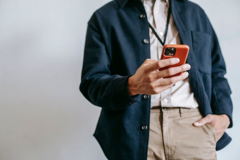 Low angle of crop unrecognizable male manager in trendy outfit and name badge messaging on mobile phone while standing near white wall with hand in pocket